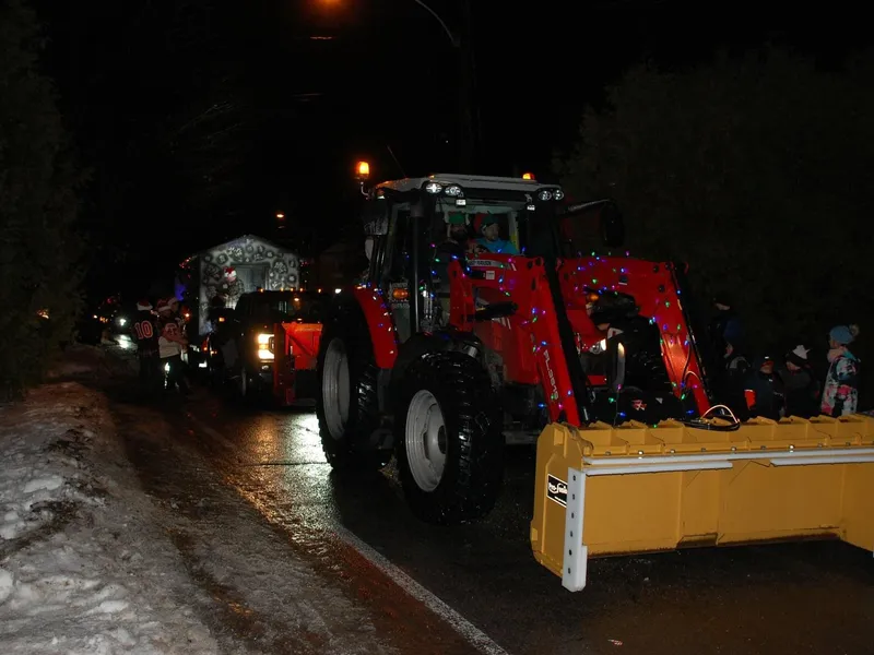 Parade de Noël de soir : le tracteur rouge de l'Entreprise SME décoré de lumières de Noël, lame jaune à l'avant, des enfants au bord de la route